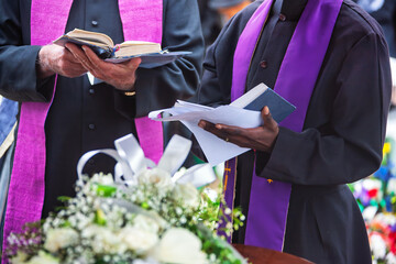 two priests reading prayers at a funeral to bless the grave at the cemetery , book the bible, coffin with flowers