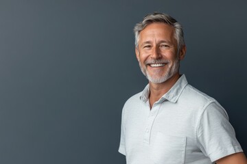 Senior male presenter in a casual shirt, smiling, standing against a solid blue-gray background with space for text