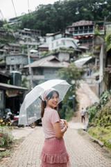 Fototapeta premium Smiling woman holding a transparent umbrella, walking down a quaint village street surrounded by lush greenery and rustic buildings.