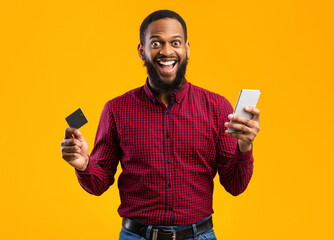 Mobile Shopping Concept. Excited African American Guy Holding Credit Card And Smartphone Posing Standing Over Yellow Background. Studio Shot. Joyful Black Man Making Online Purchase