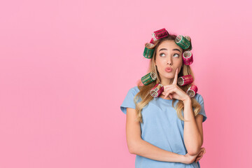Pensive young housewife in curlers presses finger to cheek and looks up to empty space, isolated on pink background, studio shot