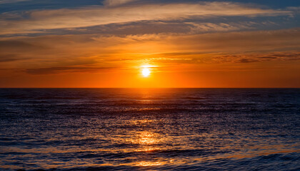 Obraz premium A tranquil sunset over the ocean, with a vibrant orange sun reflecting on the water, framed by colorful clouds.