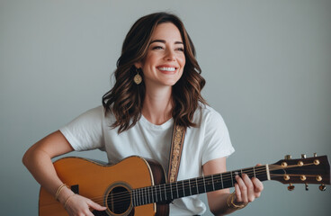smiling woman with wavy brown hair plays an acoustic guitar, radiating a relaxed and joyful mood. She wears a simple white shirt, creating a casual, warm atmosphere.