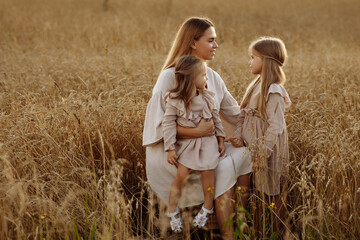 Young blonde woman with two daughters in beige dresses in a wheat field during sunset. Happy family walking.