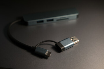 A compact, gray USB-C hub with multiple ports and attached cables, resting on a metallic surface under dim lighting. Connectivity and technology.