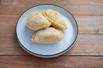Golden, flaky curry puffs rest warmly on a simple white plate, radiating a cozy, rustic charm against the wooden table backdrop.