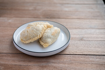 Golden, flaky curry puffs rest warmly on a simple white plate, radiating a cozy, rustic charm against the wooden table backdrop.