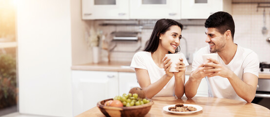 Morning Coffee. Happy couple holding mugs with hot beverage after breakfast, sitting at table, looking at each other