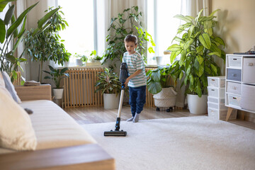 A small Caucasian European boy vacuums the carpet in the living room of the house.