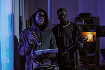 Two thieve in black clothes with crowbar, flashlight and stolen keyboard in hands hiding his face...