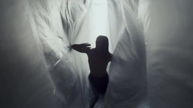 Silhouetted male dancer going towards camera with graceful movements between flowing plastic curtains, creating a dramatic, ethereal scene against backlit in studio