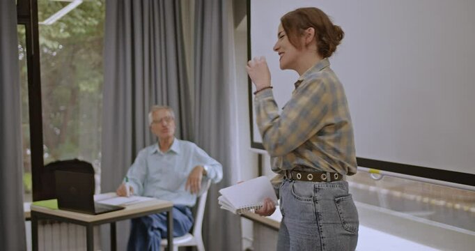 A nervous female college student stands before her class, delivering a presentation while her professor watches attentively, striving to communicate her ideas effectively despite her anxiety.