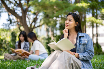 An attractive Asian woman sits on grass in a college campus park, daydreaming while reading a book.