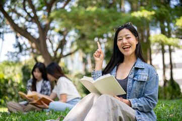 A cheerful Asian female college student sits on the grass in her college campus park.