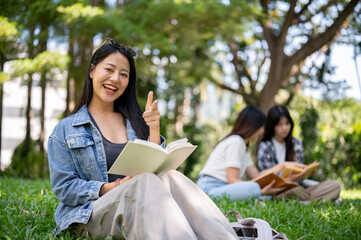 Fototapeta na wymiar A beautiful Asian female college student sits on grass in her campus park, smiling at the camera.
