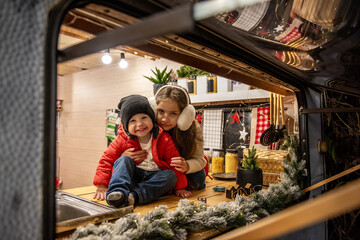 Smiling boy in a red jacket and sister in earmuffs pose in a cozy camper kitchen, decorated with Christmas lights and holiday accents. Warm family bonding during the holidays.