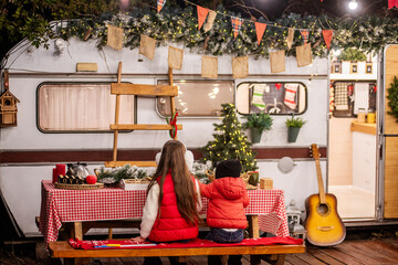 Two children in warm winter clothing sitting at a festive table outdoors, celebrating Christmas by a decorated camper van with lights and seasonal decorations.