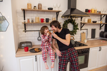 Young single father and daughter share fun moment eating cookies together while sitting on Christmas kitchen counter, surrounded by festive decor. Playful family