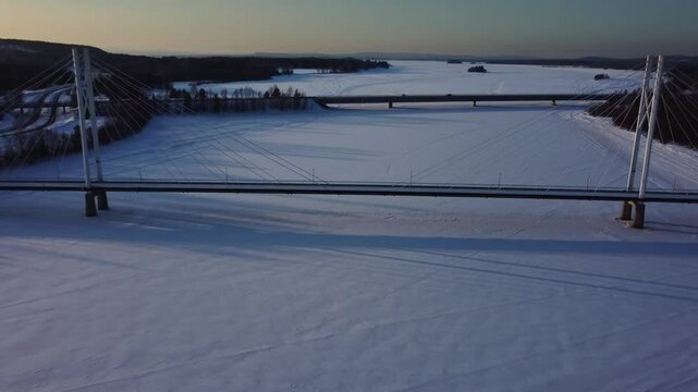 Aerial view of a cable-stayed pedestrian bridge and behind it a highway bridge with cars driving on it. Two bridges over a frozen lake during a beautiful winter sunset in nothern Sweden.