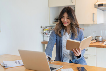 Young Freelance Woman Working from Home in Modern Kitchen, Smiling and Holding Notebook, Using Laptop for Remote Work