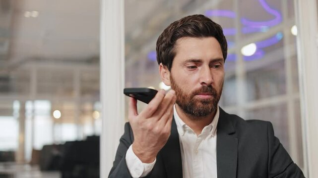 Confident office worker modern cell user hold conversation via speakerphone app record send audio message businessman sits at his desk in a modern office, talking on his phone and gesturing at laptop.