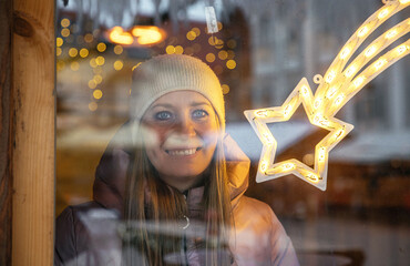 Woman in winter attire looking through a decorated Christmas window