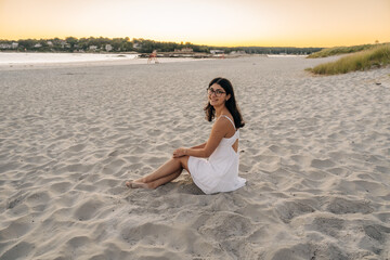 Woman sitting on sandy beach at sunset, smiling in a white dress