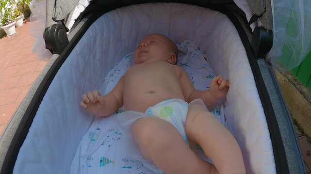 An infant is lying on its back in a pram being rolled around the backyard. The baby is wearing a patterned nappy. The baby is twirling in the pram, waving his arms and legs