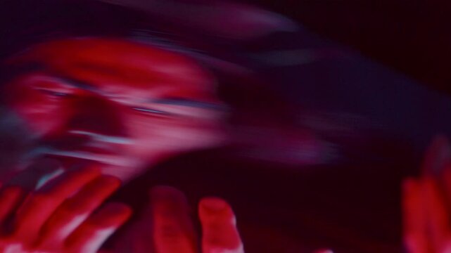 Expressive male dancer touching his face and looking at his hands in confusion in front of a distorting mirror that surreally alters his reflection in dark studio with red lighting