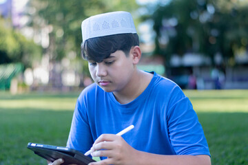 Fototapeta premium Asian muslim or islamic boy in blue t-shirt sitting on grass and writing something on his laplet, soft focus.