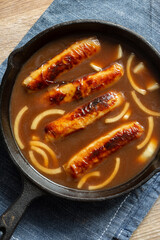 British pork sausages with onion gravy in a cast iron frying pan. On a wooden cutting board with a...