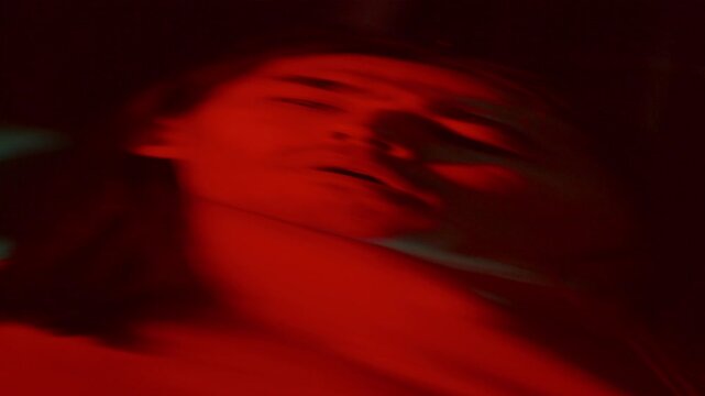 Distorted mirror reflection of long-haired man squinting and looking away in confusion in dark room with red lighting