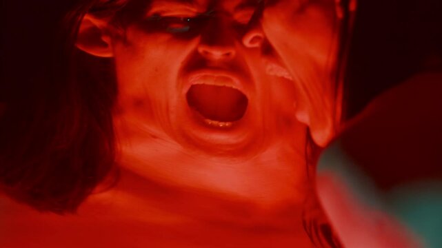 Desperate man screaming emotionally in front of distorting mirror in dark studio with red and blue lighting, agony and frustration consuming him