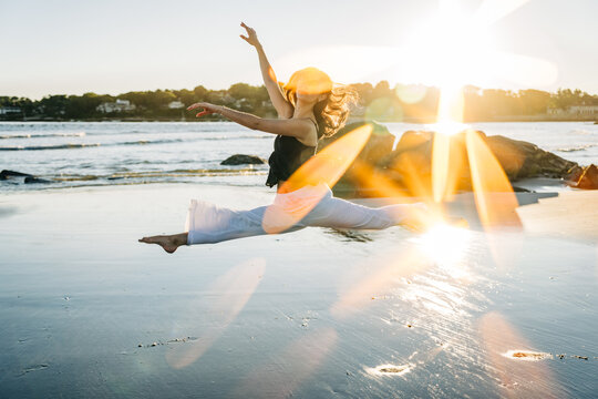 Woman performing a split leap on beach with vibrant sun flares