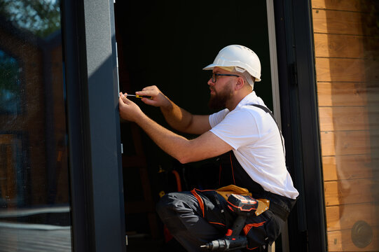 Construction worker installing door in a new house construction site. Man in working attire install front door. Builder in uniform with screwdriver adjusting front glass door in new beautiful house.