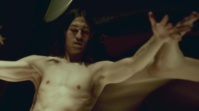 Long-haired man with shirtless torso standing captivated before large, distorting mirror that reflecting multiple versions of himself, holding arms outstretched and gazing around in awe