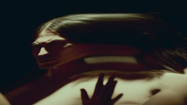 Artistic man with long hair exploring surreal, contorted reflection of himself in dimly lit room with large distorting mirror