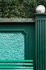 Green horizontal wooden slats of a bench against an emerald concrete fence with vertical lines and a frame surrounded by lush tropical foliage. Combination of textures and materials