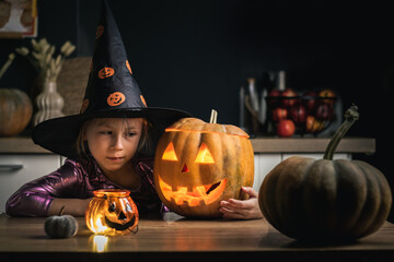 Cute girl in the spooky festive witch outfit next to creepy glowing carved Halloween pumpkin. Little girl making Jack-o-lantern while sitting at table in the kitchen. Happy Halloween background