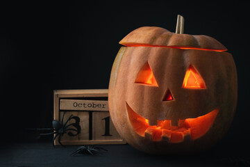 Spooky creepy glowing carved halloween pumpkin on the black background. Jack-o-lantern next to wooden block calendar with the date October 31 surrounded with spiders. Happy Halloween background