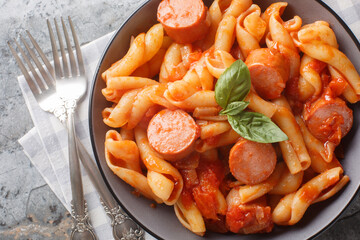 Spiral treccione pasta with smoked sausages in spicy tomato sauce close-up in bowl on table. Horizontal top view from above