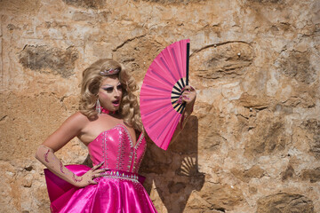 Portrait of drag queen, blonde, attractive, with crown, with pink dress, fanning herself with a big fan on a hot day, with a background of stones. Concept pride, identity, diversity, artist.