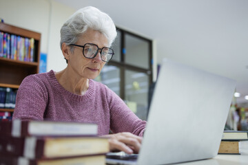 Senior woman in glasses working on a laptop in a library, surrounded by books, showcasing lifelong learning, focus, and the challenges of adapting to technology in an educational setting