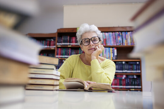 Senior woman wearing glasses in library, sitting at a desk, resting her head on her hand, looking contemplative while surrounded by books, symbolizing wisdom, lifelong learning, and reflection