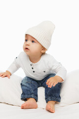Baby dressed in knitted, warm hat and white shirt, sitting on pillow and looking away against white studio background. Cozy kid's fashion. Concept of parenthood, natural beauty, love and care. Ad.