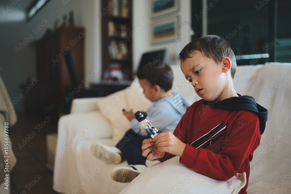 Wall mural Two young brothers, one playing with a small toy and the other using a smartphone, spend time together in their living room