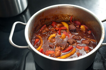Paprika goulash is braised in a stainless steel pot on a black ceramic hob, cooking at home, selected focus
