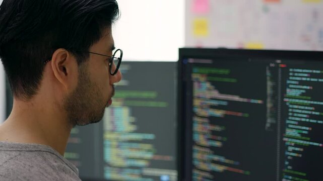Focused Asian Male Programmer Coding on Multiple Monitors in Modern Office.