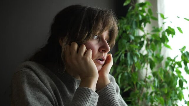Young woman is expressing sadness and worry while looking through a window. She is touching her face with different hand gestures
