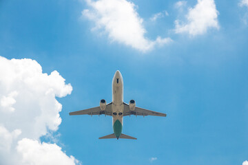 A commercial passenger plane soars through a blue sky, flanked by soft white clouds floating in the background.